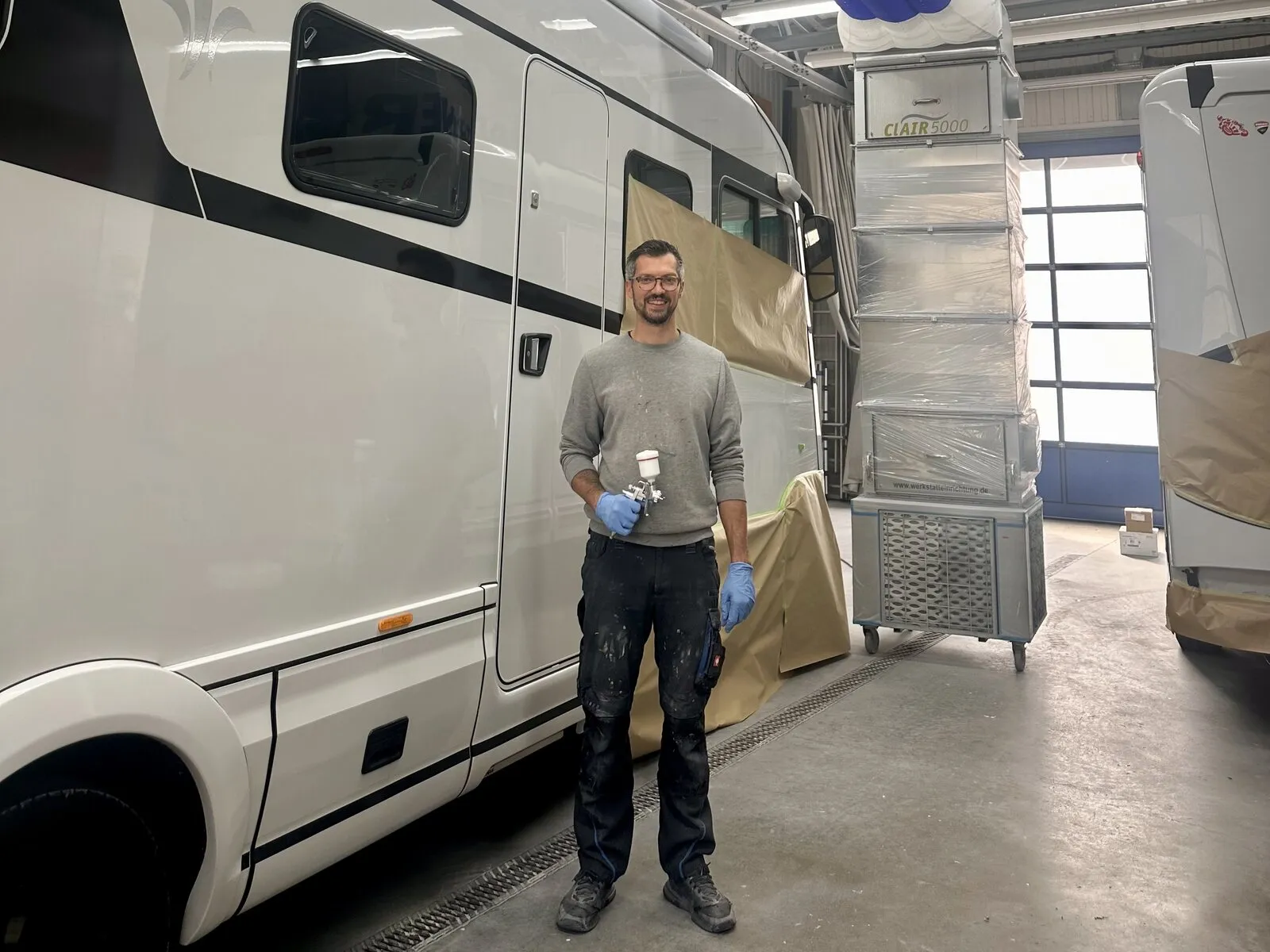 Lackierer mit Lackierpistole vor einem teilweise abgeklebten Wohnmobil in der Lackierhalle
